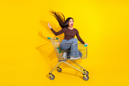 Joyful woman with flowing hair riding a shopping cart over a vibrant yellow background exuding positive energyの写真素材