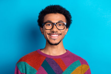 Portrait of a cheerful young man wearing glasses and a colorful sweater on a bright blue backgroundの写真素材