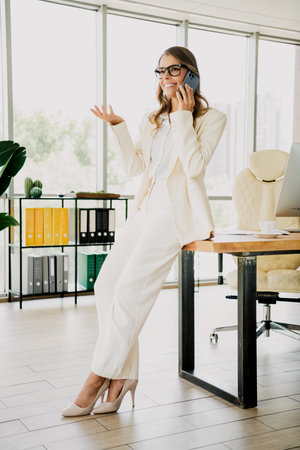 Confident young businesswoman talking on a smartphone while sitting on an office desk in a modern stylish workspaceの写真素材