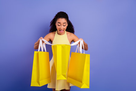 Excited young woman opening yellow shopping bags while surrounded by a vibrant purple background in a cheerful moodの写真素材