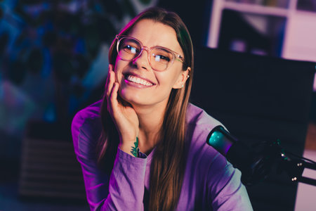 Professional female blogger with glasses in home studio broadcasting live using a modern microphone under neon lightingの写真素材