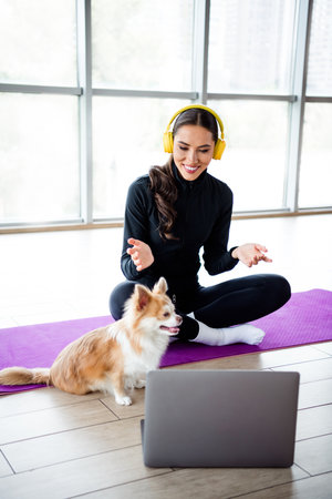 Charming woman with headphones training on yoga mat indoors while engaging with a laptop and her pet dogの写真素材