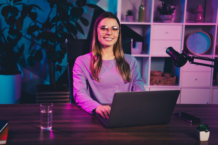 Young woman working as a media creator at home office with a laptop microphone and vibrant neon lightingの写真素材