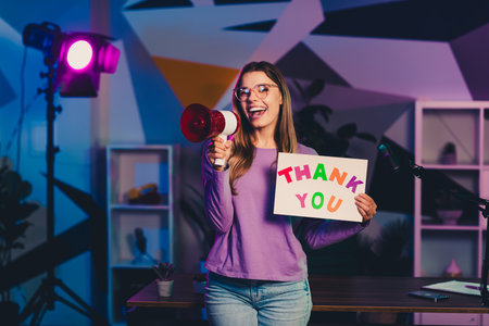 Young female blogger holding a thank you sign and megaphone in a vibrant indoor setup with neon lightingの写真素材