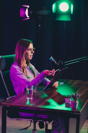 Video chat blogger podcasting in a studio with colorful lighting, utilizing a microphone setup and tablet for media content creationの写真素材
