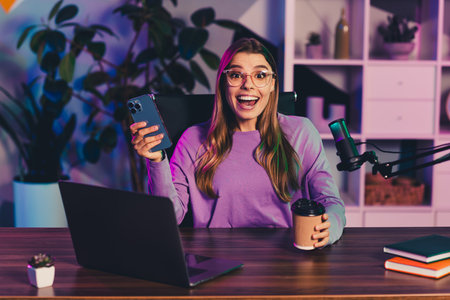 Young female blogger with laptop and phone in setup with microphone during a vibrant evening content creation sessionの写真素材