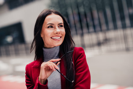 Stylish young businesswoman in a red coat outdoors confidently holding glasses while smiling on urban city streetの写真素材