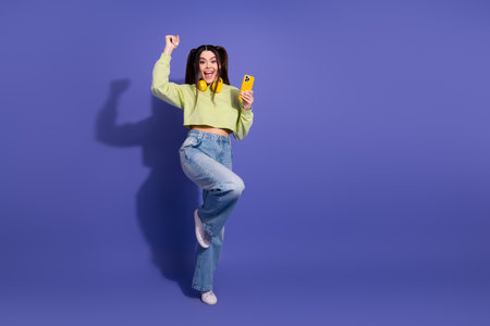Happy young woman with headphones celebrating while holding a smartphone, wearing stylish casual attire over purple backgroundの写真素材