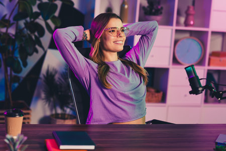 Charming woman enjoying her creative workspace with glowing neon lights and a microphone for streaming and podcastingの写真素材