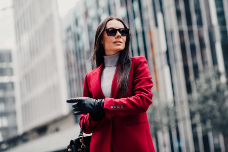 Glamorous businesswoman in a red coat walking in an urban setting during daytime, showcasing elegance and high fashionの写真素材