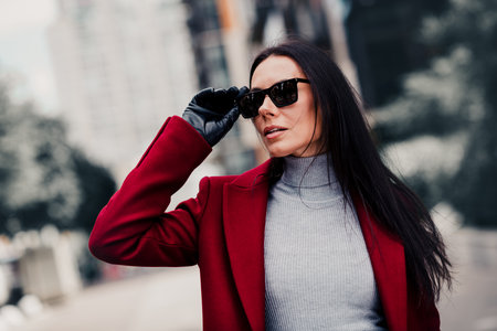 Stylish young businesswoman in a red coat and black sunglass walking outdoors in a city environment on a sunny dayの写真素材