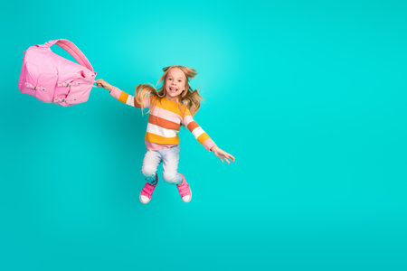 Cheerful young girl leaps high holding a pink backpack, wearing casual striped outfit, on a vibrant teal backgroundの写真素材