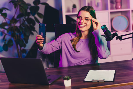 Young female podcaster recording content in a vibrant studio setting with laptop, microphone, and smartphone under colorful neon lightsの写真素材