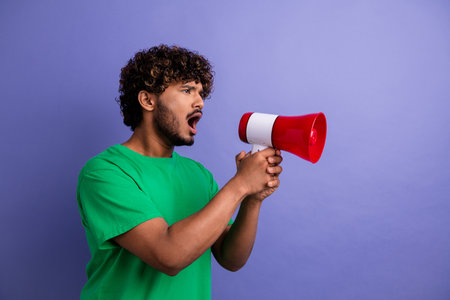 Enthusiastic Young Man Holding Red Megaphone Against Purple Wall Expressing Emotion and Making a Statementの写真素材