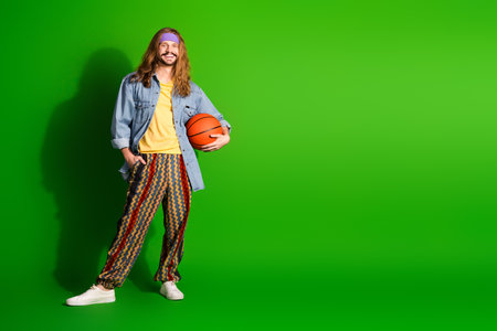 Young happy man with long hair holding basketball wearing retro outfit against green backgroundの写真素材
