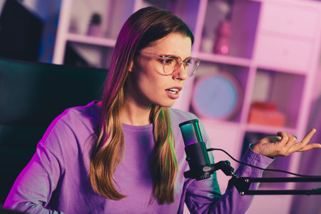 Young woman recording podcast with a microphone in a stylish room with neon lighting showcasing a creative workspaceの写真素材