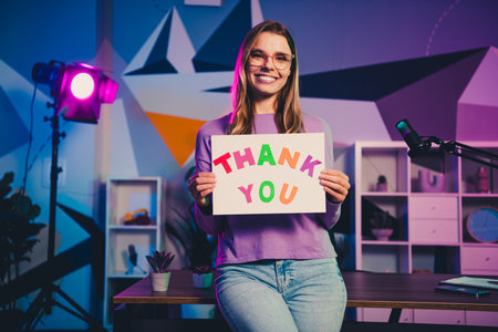 Young female blogger holding thank you sign in modern studio with vibrant lights showcasing...