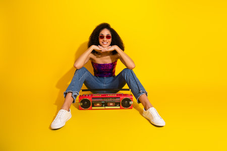Energetic young girl with curly hair wearing a purple sequin top and jeans sits against yellow...