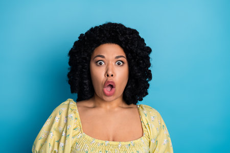 Surprised young woman with curly hair wearing yellow casual outfit in front of blue background expressing shockの写真素材