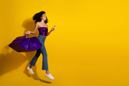 Young woman jumps with purple shopping bags smiles on her phone against a bright yellow backgroundの写真素材