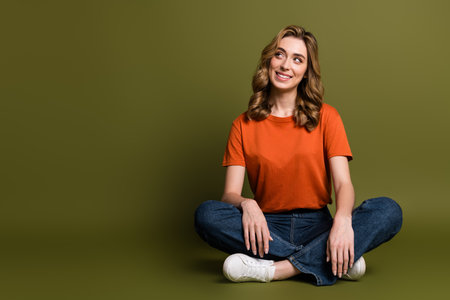 Youthful woman with stylish curls seated casually, wearing an orange t-shirt and jeans, smiling warmly on khaki backdrop.の写真素材