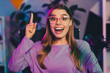 Content creator with glasses in neon-lit home studio smiling enthusiastically while live streaming or recordingの写真素材