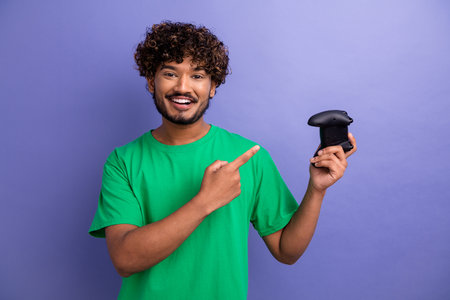 Smiling young man in a green shirt holding and pointing to a gaming controller against a simple...
