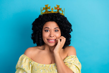 Charming young woman wearing a crown with a playful expression against a vibrant blue backgroundの写真素材