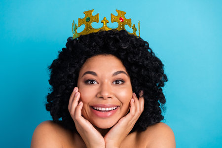 Smiling young woman wearing a crown, posing against a blue background, showcasing elegance with curly hairstyle.の写真素材
