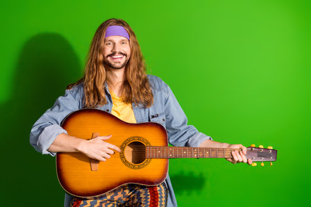 Smiling man with long hair playing acoustic guitar on a vibrant green background, embodying a retro bohemian styleの写真素材