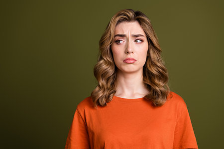 Young thoughtful woman in a casual orange shirt with wavy blonde hair standing against a khaki backgroundの写真素材