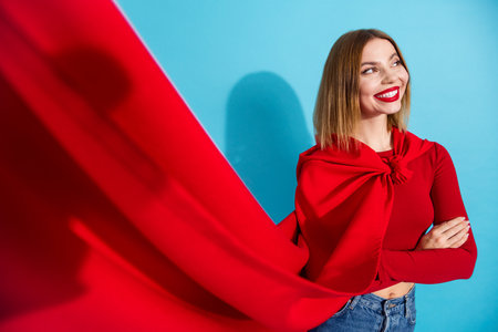Charming young woman in vibrant red outfit smiling against a bright blue background in a stylish and cheerful poseの写真素材
