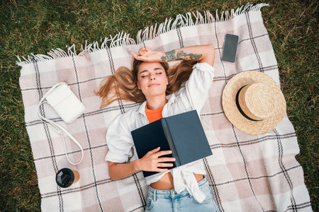 Young woman relaxing outdoors in a sunny park reading a book on a plaid blanket with a straw hat nearby a phone coffee cup and a hopeful mood during a bright summer dayの写真素材