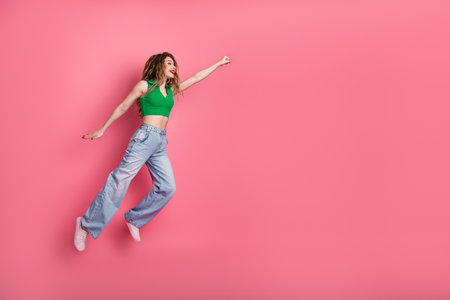Woman in green crop top and jeans smiling and jumping playfully against pink backgroundの写真素材