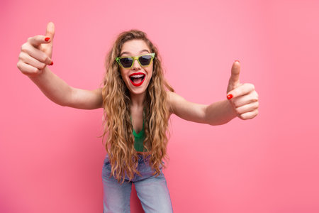 Young woman in green top and sunglass giving dynamic thumbs up gesture against vibrant pink backgroundの写真素材