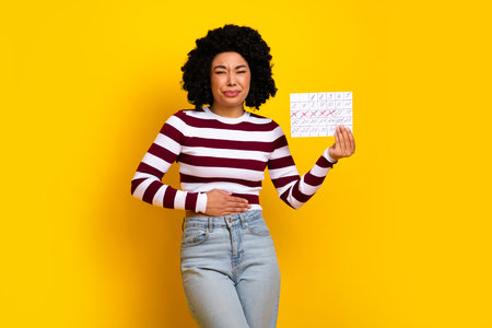 Young woman holding calendar, expressing discomfort, wearing trendy striped pullover on yellow backgroundの写真素材