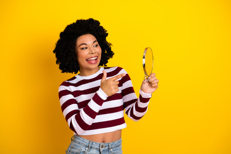 Cheerful stylish woman holding a round mirror in her hand against a vibrant yellow background with a captivating smileの写真素材