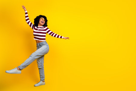 Cheerful young woman with curly hair in a casual striped top dancing joyfully against a vibrant yellow backgroundの写真素材