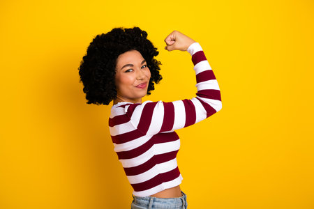 Confident young woman showing strength in striped top on vibrant yellow background, embodying empowerment and styleの写真素材