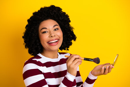Cheerful woman applying makeup with a brush against a vibrant yellow background, showcasing beauty and positivityの写真素材