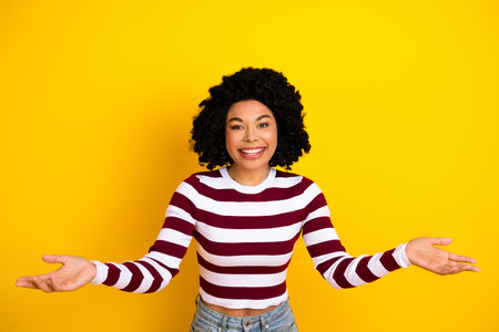 Happy young woman with curly black hair posing casually in striped top on vibrant yellow background,...