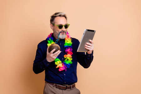Businessman Enjoys Leisure Time Using Tablet While Holding Coconut and Wearing Colorful Lei on Beige Backgroundの写真素材