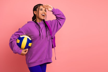 Youthful woman in vibrant purple sportswear holding volleyball and gesturing with a cheerful smile against pink backgroundの写真素材