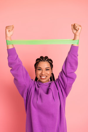 Young woman in a purple hoodie smiling and exercising with a green resistance band against a pink...