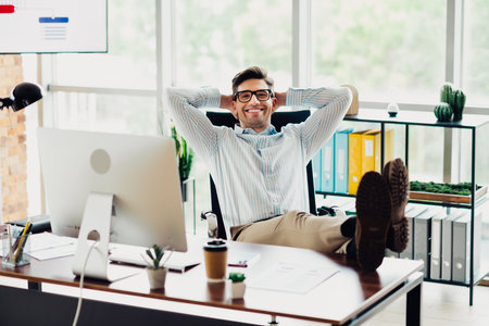 Confident young businessman relaxing at his modern office desk with a smile, showcasing a successful and stylish workspace.の写真素材