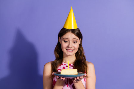 Young woman in a party hat holding a plate with cake and candle against vibrant colorful background.の写真素材