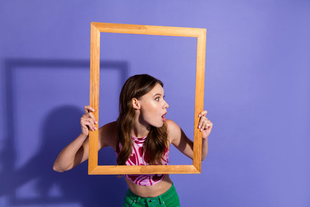 Young stylish woman with a playful expression holding a decorative wooden frame against a vibrant purple backgroundの写真素材