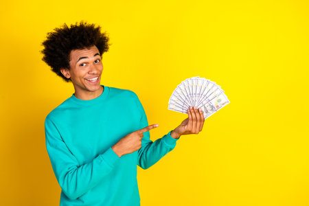 Smiling young man with curly hair holding and pointing at a fan of dollar bills on a bright yellow backgroundの写真素材