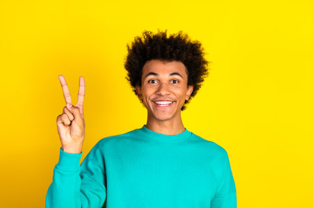 Young man with a joyful smile showing a peace gesture against vibrant yellow background wearing a teal sweatshirtの写真素材