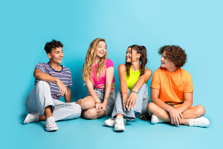 Four friends teens sit together on a bright blue studio backdrop wearing colorful casual outfits and...